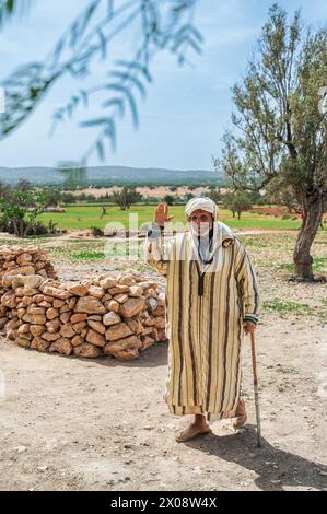 A senior man dressed in a traditional Moroccan jellaba waves at the camera, standing in a rural Moroccan landscape with a walking stick Stock Photo