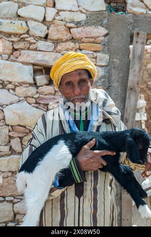 An elderly Moroccan man, wearing a traditional yellow turban, holds a young black and white goat affectionately in front of a stone wall Stock Photo