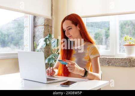 A redheaded woman with a serene smile is seen making an online booking for a nature getaway, holding a credit card while using her laptop in a room wi Stock Photo