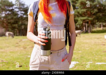 Close-up of anonymous redheaded woman holding a water bottle, dressed for hiking in nature, with trees and a meadow in the background Stock Photo
