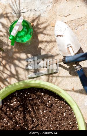 Gardening tools arranged on a patio including a green watering can, a spade, and a hand fork, with a pot filled with soil ready for planting. Stock Photo