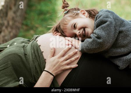 A sweet moment as a young girl with a big smile listens to a pregnant belly, bonding with the unborn sibling outdoors Stock Photo