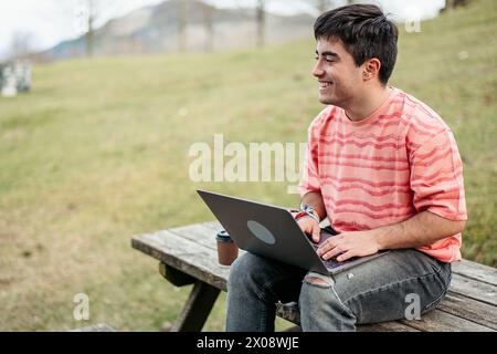 A man is deeply engrossed in his work on a laptop while seated alone at a wooden picnic table, surrounded by the tranquility of nature Stock Photo