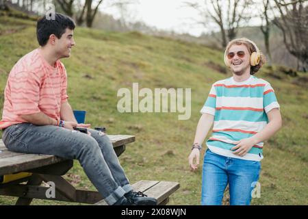Two happy young men share a laugh while enjoying the outdoors, with one sitting at a park bench and the other standing, conveying a relaxed and joyful Stock Photo
