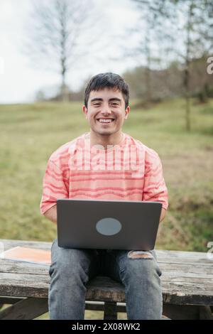 A happy young man works on his laptop in the park, with a bright smile, enjoying the flexibility of outdoor remote work Stock Photo