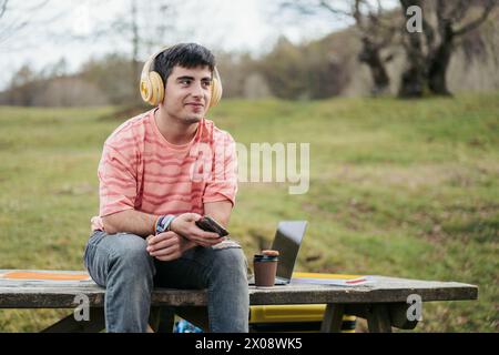 A relaxed young man sits on a park bench with headphones, a smartphone in hand, and a laptop on the table, embodying a casual work-from-anywhere vibe Stock Photo