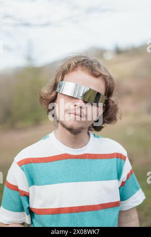 A young man poses in unique, mirrored sunglasses, exuding a cool and contemporary vibe in a natural outdoor setting Stock Photo