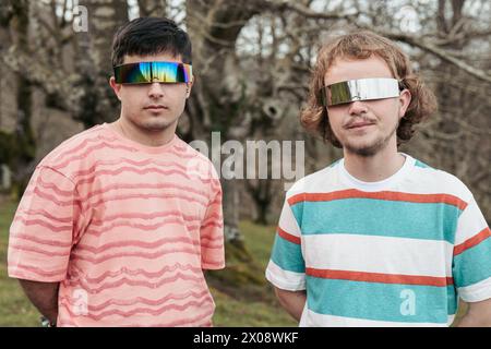 Two men stand side by side, exuding confidence and style in their bold mirrored sunglasses, set against a natural outdoor backdrop Stock Photo