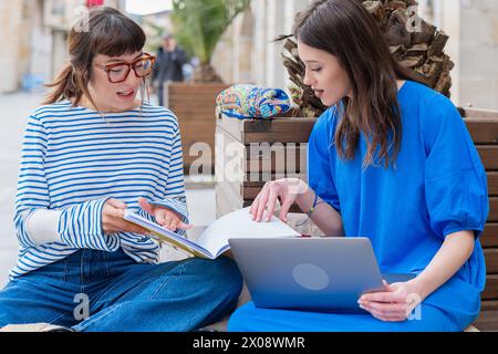 A pair of women are engaged in work or study on a bench outdoors, with one on a laptop and the other taking notes, both wearing bright spring attire Stock Photo