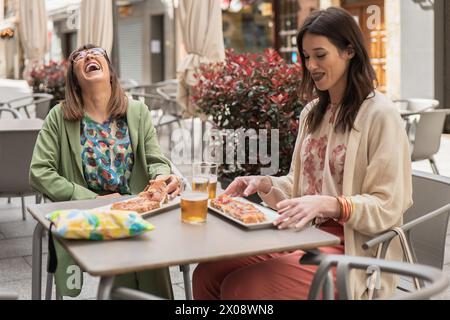Two joyful women sharing a moment of laughter while eating pizza and having drinks at an outdoor cafe, highlighted by springtime attire Stock Photo