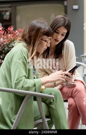 A pair of fashionable women engaged in using a smartphone, dressed in light, spring-themed clothing, showcase a casual and connected lifestyle Stock Photo