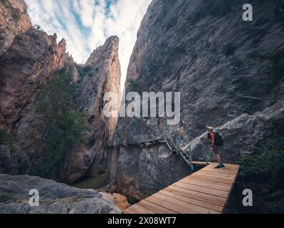 An intrepid hiker pauses on a wooden boardwalk, admiring the sheer rock faces of a narrow gorge, under a serene blue sky Stock Photo