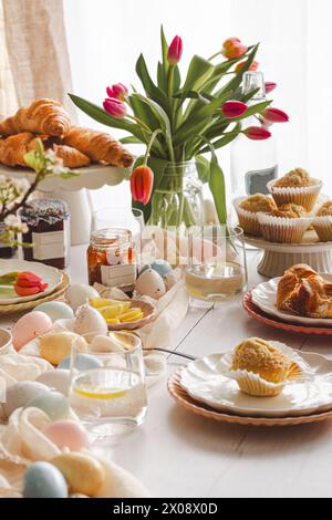 An inviting Easter breakfast table featuring fresh croissants, coconut muffins, marmalade jars, and a bouquet of tulips among colorful eggs and tablew Stock Photo