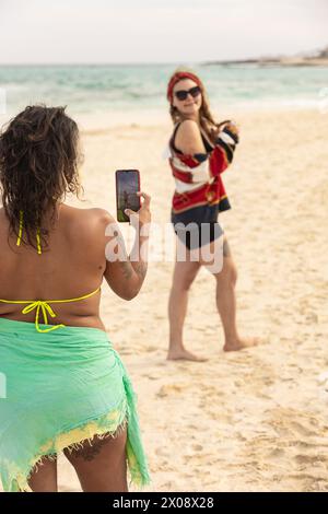 A joyful LGBTQI female couple is captured having a playful moment on a sandy beach, with one woman taking a photo of the other who is posing joyfully. Stock Photo