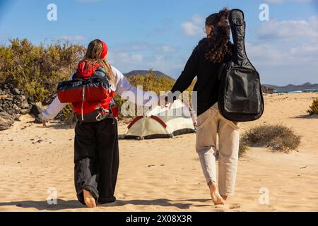 A back view loving couple walks hand in hand near their campsite on the sandy beach, creating memories with a mix of adventure and music Stock Photo