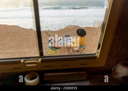 Back view of unrecognizable nomadic male musician composing music by the sea at sunset, viewed from inside a motorhome with a laptop and guitar. Stock Photo