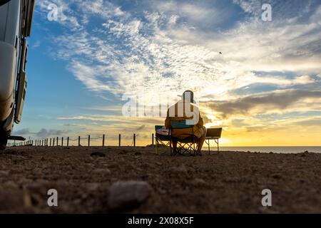 Back view of unrecognizable male musician sits by his motorhome, composing on his laptop, inspired by the coastal sunset. Stock Photo