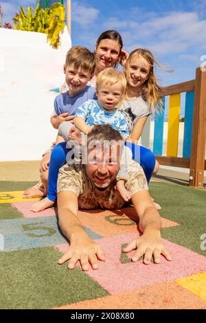 A happy family enjoys playtime at a playground, with a smiling father lying on the ground, mother and children piled on his back, including a baby wit Stock Photo