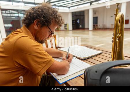 Side view of a man with one arm writes music while sitting next to his trumpet in a well-lit home courtyard, illustrating artistic determination. Stock Photo