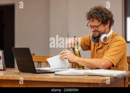 A focused one-armed trumpeter composes music in his home courtyard, using a laptop and writing notes on sheet music. Stock Photo