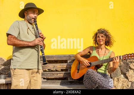 A street music duo performs on a sunny day, with a woman playing guitar and a man playing clarinet against a vivid yellow wall. Stock Photo