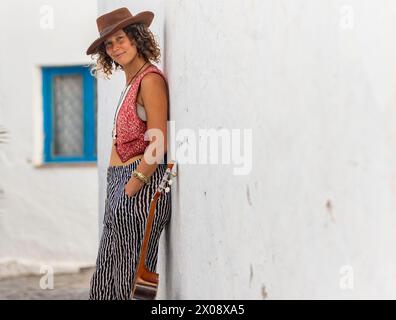 A female street musician leaning against a white wall, holding a guitar, in a charming town setting. Stock Photo
