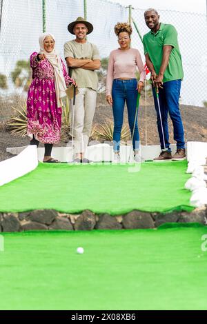 A group of four diverse friends share laughter while playing mini golf on a lush green course. Stock Photo