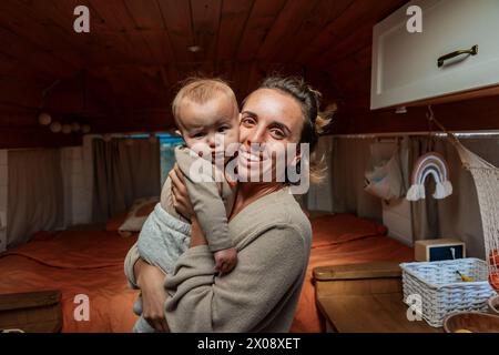 Smiling woman holding a baby inside a cozy, wood-paneled van converted into a family custom home Stock Photo