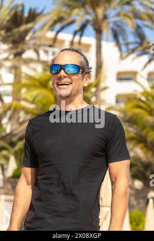 A cheerful man wearing sunglasses and a black shirt radiates happiness while standing in a tropical resort setting with palm trees in the background. Stock Photo