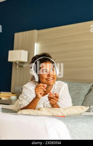 A smiling middle aged hispanic woman enjoys listening to music with headphones, comfortably lounging on a couch at home, exuding casual relaxation and Stock Photo