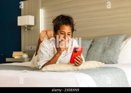 A smiling middle aged hispanic woman enjoys leisure time while browsing on her smartphone, lying comfortably on a hotel bed with stylish interior deco Stock Photo