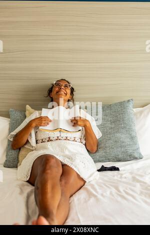 Cheerful middle aged hispanic woman in glasses lying on a bed, enjoying a book in a comfortable bedroom with stylish pillows and soft lighting. Stock Photo