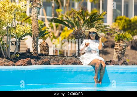 A smiling middle aged hispanic woman in a white dress and sunhat sits at the edge of a pool, holding a cocktail, surrounded by exotic plants. Stock Photo