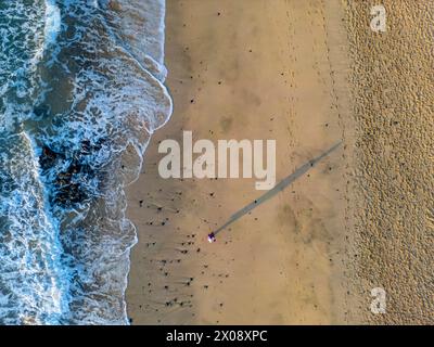 An aerial shot captures a single woman standing on a sandy beach, the waves gently reaching toward her Stock Photo