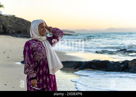 A joyful Muslim woman in a hijab stands on a sandy beach, smiling as she experiences the tranquil beauty of a seaside sunset Stock Photo