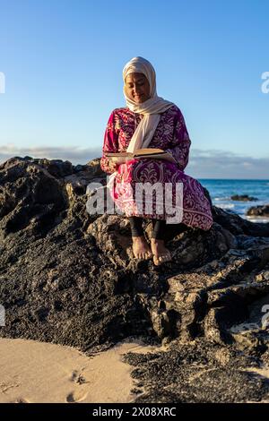 A Muslim woman in a colorful dress and white hijab sits on dark rocks, contemplating as the sunset paints the sky Stock Photo