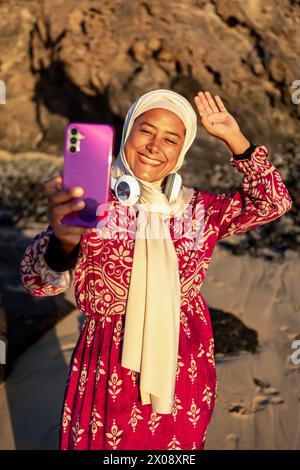 A joyful Muslim woman in a hijab is taking a selfie on the beach, waving with one hand, holding a pink smartphone Stock Photo