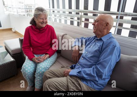 An elderly man and woman laugh together while sitting on a comfortable sofa, showcasing warmth and companionship. Stock Photo