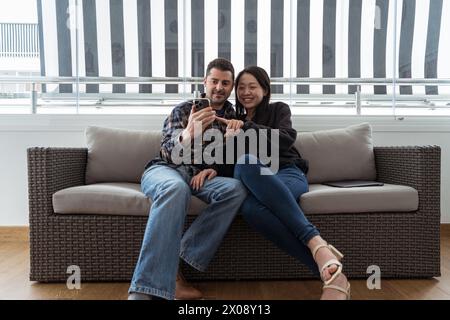 Young multiethnic couple enjoys a cozy moment on a sofa, using a smartphone together with smiles and casual clothing. Stock Photo