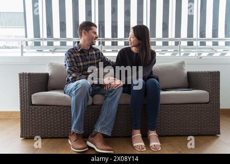 An multiethnic happy couple sits closely on a sofa, engaging in a conversation with joyful expressions. Stock Photo