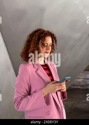 A curly-haired young redhead woman dressed in a stylish pink blazer is focused on her smartphone Stock Photo