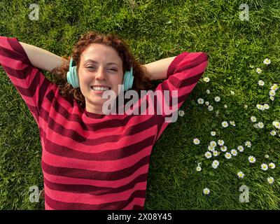 A cheerful young redhead woman lies on the grass with her arms stretched out, enjoying music on her wireless headphones, surrounded by daisies Stock Photo