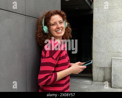 A joyful young redhead woman with curly hair smiles as she listens to music on teal headphones, holding a smartphone Stock Photo