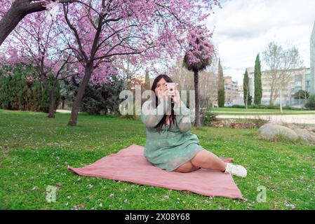 A woman sits on a pink blanket in the park, capturing photos with a camera under blooming cherry blossom trees. Stock Photo