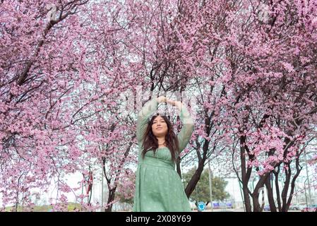 A woman in a green dress with her hands up among pink cherry blossoms. Stock Photo