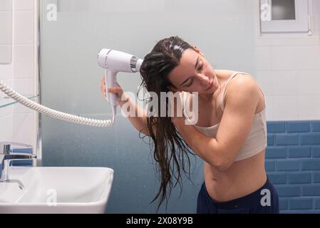 A young woman in casual clothing blow-drying her wet hair in a tiled bathroom after shower time, with a serene expression Stock Photo