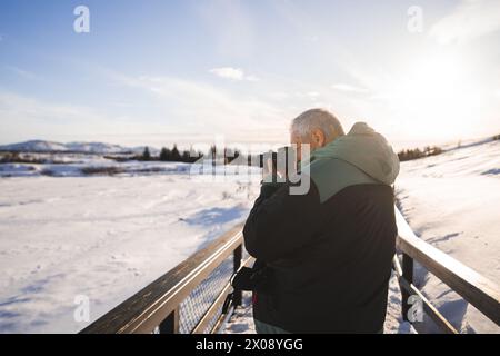 A photographer stands on a bridge in Iceland, camera in hand, capturing the stunning winter scenery bathed in the soft glow of the sun Stock Photo