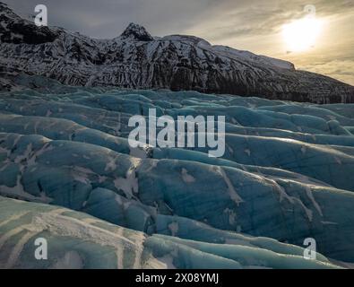 Sun gently illuminates the vast blue crevasses of an Icelandic glacier with snow-covered mountains in the background Stock Photo