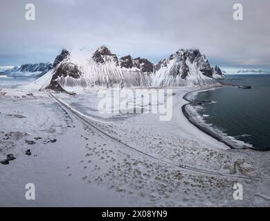 Panoramic view of the snow-covered mountains beside a tranquil coastline in Iceland, under a cloudy sky Stock Photo