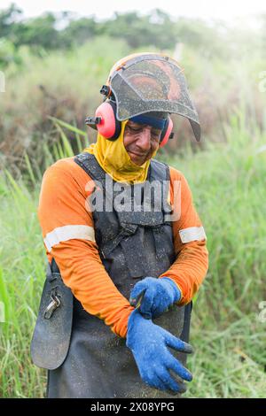 A farmer stands in a lush field, his safety helmet with a visor and earmuffs on, he's taking a break from using a scythe, wearing a reflective vest an Stock Photo